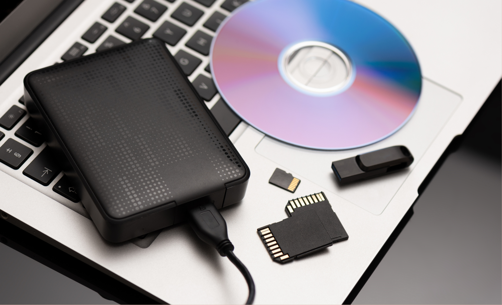 External storage devices including a portable hard drive, USB flash drive, SD cards, and a DVD resting on a laptop keyboard to free up disk space